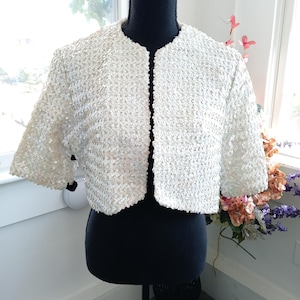 Puede incluir: Una chaqueta bolero blanca de lentejuelas con mangas cortas. La chaqueta está en un maniquí negro.