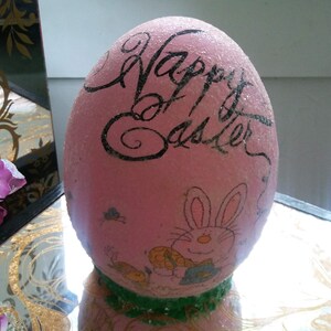 May include: A pink Easter egg with glitter, decorated with the words "Happy Easter" and a cartoon bunny. The egg is sitting on a table with a gold and black marbled surface.