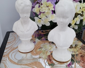 May include: Two white ceramic busts of people, one with short hair and the other with a bun. The busts are on a mirrored surface with a floral design, and a bouquet of flowers is in the background. The busts are likely decorative items.