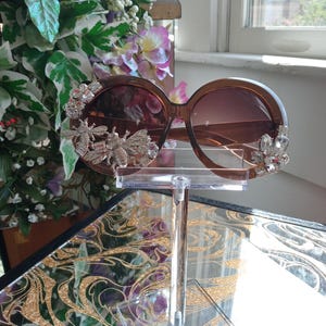 Puede incluir: Gafas de sol marrones de gran tamaño con detalles decorativos de abeja plateada y cristal. Las gafas de sol se exhiben sobre un soporte transparente, con un arreglo floral en el fondo. Las lentes tienen un tinte degradado.