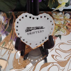 May include: A heart-shaped white ceramic dish with a gold rim and heart-shaped cutouts. The dish features the word "DRIFTERS" in black text and a black graphic. The dish is displayed on a dark brown stand.