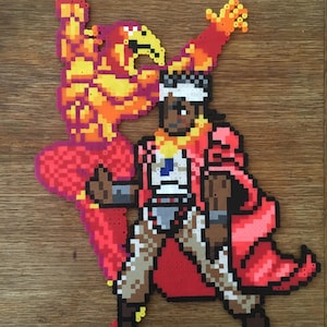 May include: A pixel art figure of a person in a red cape and white head covering, with a matching red and yellow figure behind them. The figure is made of small, colorful beads, and the background is a wooden surface.