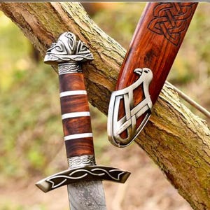 Puede incluir: Espada vikinga con empuñadura de madera y detalles metálicos plateados. La hoja presenta un diseño. La vaina de madera muestra un diseño de nudo celta. La espada se apoya sobre una rama.