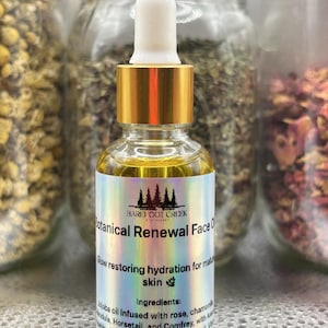 Op de afbeelding: Een heldere glazen fles Botanical Renewal Face Oil met een gouden druppelkap. Het etiket bevat de tekst "Barefoot Creek" en "Glow restoring hydration for mature skin". De achtergrond bevat potten met gedroogde kruiden.