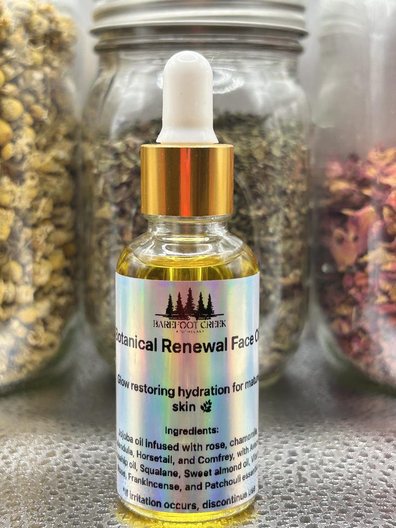Op de afbeelding: Een heldere glazen fles Botanical Renewal Face Oil met een gouden druppelkap. Het etiket bevat de tekst "Barefoot Creek" en "Glow restoring hydration for mature skin". De achtergrond bevat potten met gedroogde kruiden.