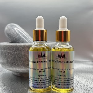 Op de afbeelding: Twee glazen flessen Barefoot Creek Botanical Renewal Face Oil met gouden doppen en pipetten. De flessen bevatten een gele vloeistof en hebben etiketten met tekst. Een vijzel en stamper staan op de achtergrond.