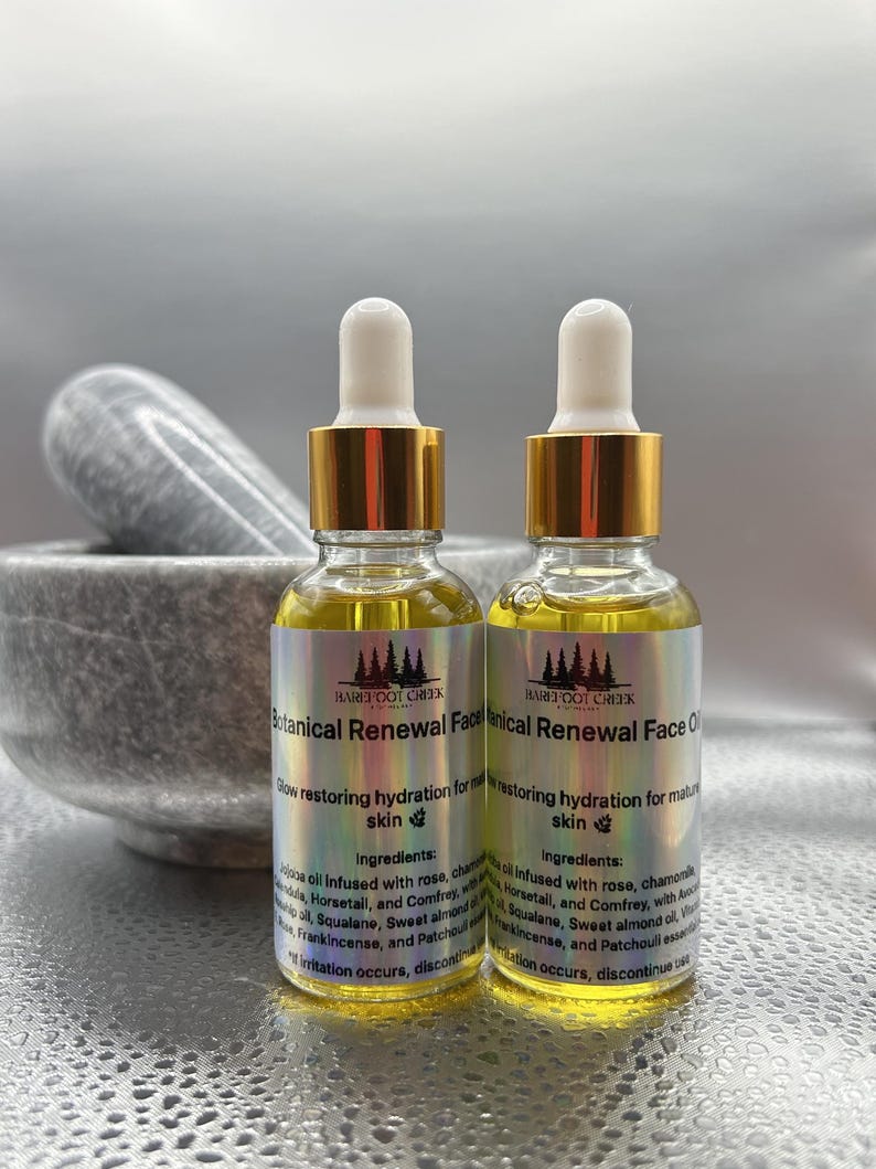 Op de afbeelding: Twee glazen flessen Barefoot Creek Botanical Renewal Face Oil met gouden doppen en pipetten. De flessen bevatten een gele vloeistof en hebben etiketten met tekst. Een vijzel en stamper staan op de achtergrond.