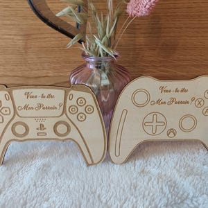 Peut inclure: Deux découpes en bois en forme de manettes de jeu, une PlayStation et une Xbox. Les deux portent l'inscription gravée "Veux-tu être Mon Parrain ?" en écriture cursive. Les manettes sont de couleur marron clair et reposent sur une surface blanche texturée.