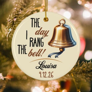 May include: A round, cream-colored ceramic ornament featuring a gold-toned bell illustration and the text "THE day I RANG THE bell!" along with the name "Louisa" and the date "9.12.26" in black and brown lettering, suspended by a gold string.