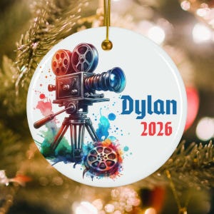 May include: A round white ceramic ornament featuring a colorful watercolor illustration of a vintage film camera on a tripod with a film reel at the base. The text Dylan is printed in blue and the year 2026 is printed in red on the right side.