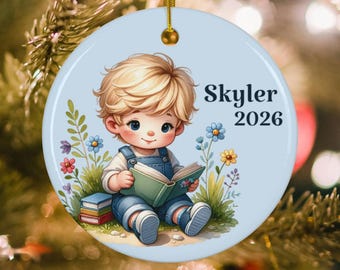 May include: A round ceramic ornament with a light blue background featuring a blonde child in denim overalls reading a green book while sitting in grass with colorful flowers. A stack of books is nearby. The text Skyler 2026 is printed in black.