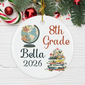 May include: A round white ceramic ornament featuring a gold hanging string. The design includes a globe with floral accents, a stack of books topped with flowers, and the text 8th Grade, Bella, and 2026 printed in dark brown and black fonts.