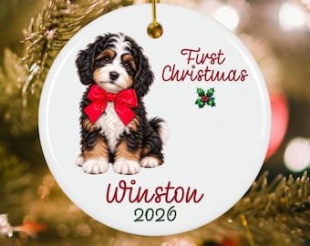 May include: A round white ceramic ornament featuring a black, white, and brown puppy wearing a red bow. The text reads First Christmas, Winston, and 2026, accompanied by a small green holly illustration with red berries and a gold hanging string.