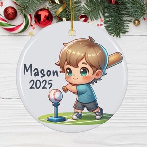 May include: A white ceramic Christmas ornament featuring a cartoon illustration of a boy playing baseball. The boy is wearing a blue shirt and has brown hair. The ornament is personalized with the name "Mason" and the year "2025".