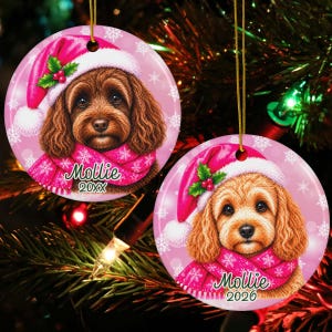 May include: Two round ceramic Christmas ornaments featuring a brown curly-haired dog wearing a pink Santa hat with holly and a matching pink scarf. The ornaments are printed with the name Mollie and the years 20XX and 2026 on a pink background.