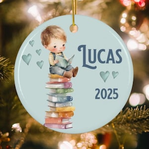May include: A blue and white personalized Christmas ornament featuring a cartoon boy reading a book on a stack of books. The ornament says "Lucas 2025" and has small blue hearts around the edges.