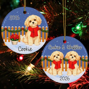 May include: Two round ceramic ornaments featuring illustrations of cream-colored dogs wearing red scarves. One ornament shows a single dog with the text 2026 and Cookie. The other shows two dogs with the text Cookie & Waffles and 2026.