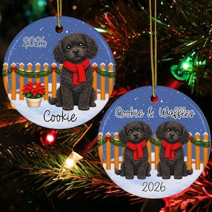 May include: Two round ceramic ornaments featuring illustrations of black curly-haired dogs wearing red scarves. One ornament shows a single dog with a poinsettia and the text 2026 and Cookie. The other shows two dogs with the text Cookie & Waffles and 2026.