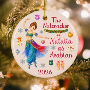 May include: A round ceramic ornament featuring a dancer in a blue and red Arabian-style costume. The text reads The Nutcracker Natalia as Arabian 2026. The design includes illustrations of snowflakes, bows, ornaments, and ballet slippers.