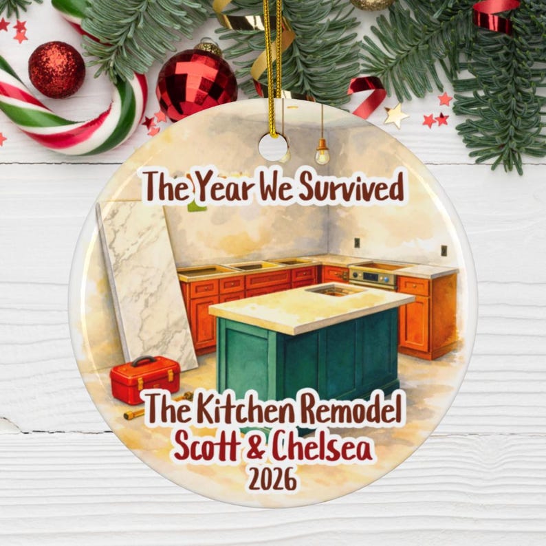 May include: A round ceramic ornament featuring a watercolor illustration of a kitchen remodel with orange cabinets, a green island, and a red toolbox. The text reads: The Year We Survived, The Kitchen Remodel, Scott & Chelsea, 2026. It has a gold string.