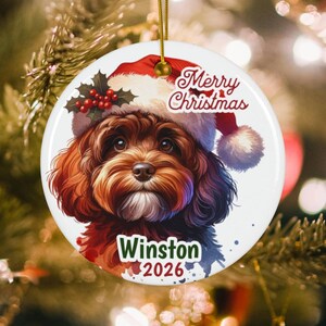 May include: A round white ceramic Christmas ornament featuring a brown curly-haired dog wearing a red Santa hat with holly berries. The text Merry Christmas is printed at the top, and the name Winston and the year 2026 are printed at the bottom.