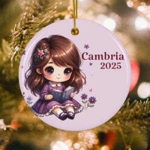 May include: A round, light purple ceramic ornament with a cartoon illustration of a girl reading a book. The girl has brown hair and wears a purple dress. The ornament has the name "Cambria" and the year "2025" printed on it.