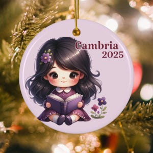 May include: A round, light purple ceramic ornament with a cartoon girl reading a book. The girl has dark hair and wears a purple dress. The ornament has the text "Cambria 2025" in a decorative font. A gold string is attached for hanging.