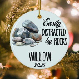 May include: A round white ceramic ornament featuring a watercolor illustration of a pile of grey and brown rocks. The text reads "Easily DISTRACTED by ROCKS" in black script and print, followed by the name "WILLOW" and the year "2026" in black.