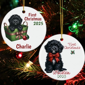 May include: Two white ceramic Christmas ornaments with gold hanging strings. One features a black puppy in a green basket with a red bow, the text "First Christmas 2025" and the name "Charlie". The other shows a black puppy with a red bow, the text "First Christmas 2025" and the name "Winston".