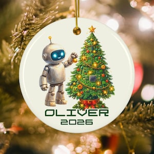 May include: A round white ceramic ornament featuring a silver robot with glowing blue eyes decorating a green Christmas tree made of circuit boards. The tree is topped with a gold star. The text OLIVER 2026 is printed in green below the image.