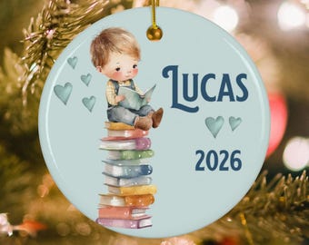 May include: A round, light blue ceramic ornament featuring a watercolor illustration of a young child reading a book while sitting on a stack of colorful books. The text "LUCAS" and "2026" are printed in dark blue, accompanied by small heart icons.