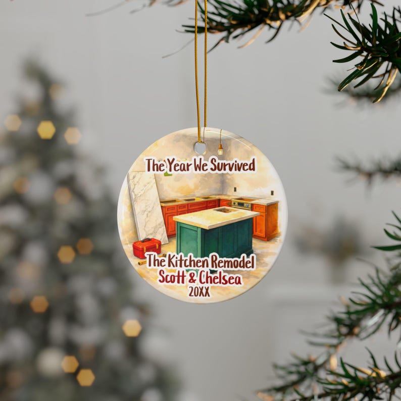 May include: A round ceramic ornament hanging by a gold string, featuring an illustration of a kitchen remodel with a green island and orange cabinets. The text reads: The Year We Survived The Kitchen Remodel Scott & Chelsea 20XX.