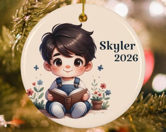 May include: A round ceramic ornament featuring a cartoon illustration of a child with dark hair wearing blue overalls and reading a book. The design includes small colorful flowers and the text Skyler 2026. A gold string is attached to the top.