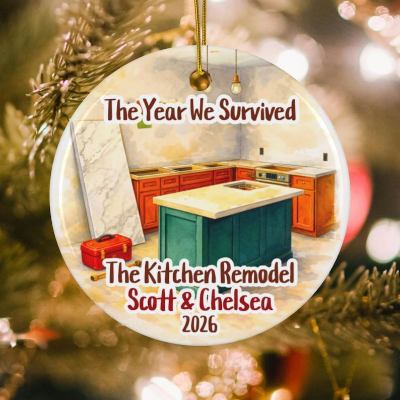 May include: A round ceramic ornament featuring an illustration of a kitchen remodel with orange cabinets and a green island. The text reads The Year We Survived The Kitchen Remodel Scott & Chelsea 2026. A gold string is attached to the top for hanging.