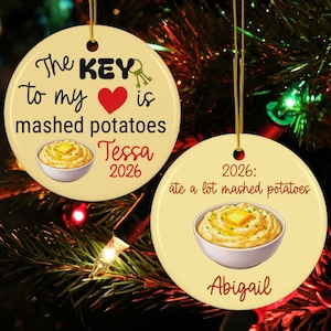 May include: Two round, cream-colored ceramic ornaments with gold hanging strings. One features the text 'The KEY to my heart is mashed potatoes', a red heart, a bowl of mashed potatoes with butter, 'Tessa', and '2026'. The other says '2026: ate a lot mashed potatoes' with a bowl of mashed potatoes and 'Abigail'.