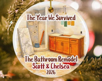 May include: A round ceramic ornament featuring an illustration of a bathroom under construction with wooden cabinets, a marble slab, and tools. The text reads: The Year We Survived, The Bathroom Remodel, Scott & Chelsea, 2026. It has a gold string.
