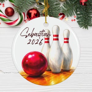 May include: A round white ceramic ornament featuring a glossy red bowling ball and three white bowling pins with red stripes. The text "Sebastian" and "2026" is printed in black script and block lettering. A gold string is attached at the top.