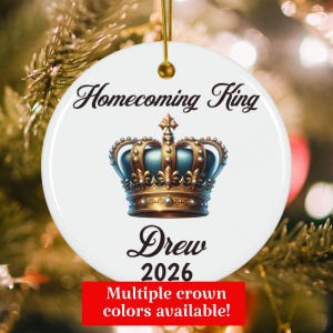 May include: A round white ceramic ornament featuring a gold crown graphic in the center. The text "Homecoming King" is printed in black script above the crown, with the name "Drew" and the year "2026" printed in black below the crown.