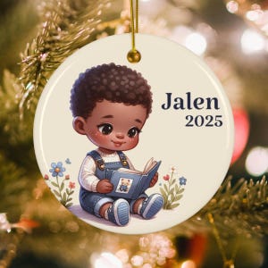 May include: A round, cream-colored ceramic ornament featuring a cartoon illustration of a child reading a book. The child is wearing denim overalls and a white shirt. The name "Jalen" and the year "2025" are printed on the ornament.
