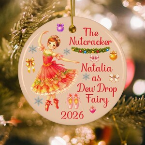 May include: A round ceramic Christmas ornament featuring a ballerina in a red and yellow dress, ballet slippers, and snowflakes. The text reads The Nutcracker, Natalia as Dew Drop Fairy, and 2026. It includes a gold hanging string and loop.