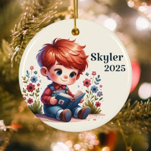 May include: Round ceramic ornament featuring a cartoon illustration of a child with red hair reading a book. The ornament has the name "Skyler" and the year "2025" printed on it. The background is decorated with flowers and greenery.