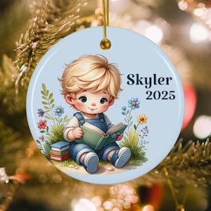 May include: A round, light blue Christmas ornament featuring a cartoon illustration of a child reading a book. The child is wearing denim overalls and surrounded by flowers. The name "Skyler" and the year "2025" are printed on the ornament.
