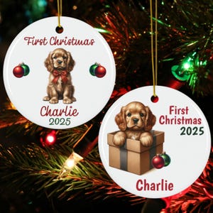 May include: Two white ceramic Christmas ornaments with gold hanging strings. One features a puppy with a red bow, the text "First Christmas", "Charlie", and "2025". The other shows a puppy in a box with the same text. Red and green ornaments are also present.