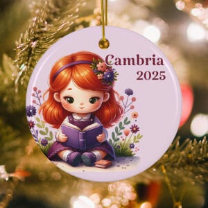 May include: A round, light purple Christmas ornament with a cartoon illustration of a girl reading a book. The girl has red hair and is wearing a purple dress. The text "Cambria 2025" is printed on the ornament.