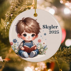 May include: Round ceramic ornament featuring a cartoon boy reading a book, surrounded by flowers. The ornament has the name "Skyler" and the year "2025" printed on it. The ornament is hanging from a gold string.