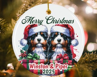 May include: A round white ceramic Christmas ornament featuring two Bernese Mountain Dogs wearing red Santa hats. Below the dogs are two gift boxes with red and green bows. The text reads Merry Christmas, Winston & Piper, and 2025.