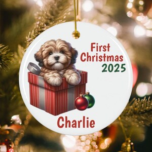 May include: A round white ceramic Christmas ornament featuring a fluffy brown puppy sitting inside a red and white striped gift box. The text reads First Christmas 2025 and the name Charlie in red and green. A gold string is attached at the top.