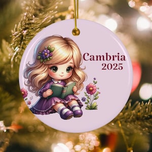 May include: A round, light purple Christmas ornament featuring a cartoon girl with blonde hair, wearing a purple dress, and reading a book. The ornament has the text "Cambria 2025" in brown.