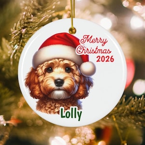 May include: A round white ceramic ornament featuring a fluffy brown puppy wearing a red Santa hat. The text Merry Christmas 2026 is printed in red script above the puppy, and the name Lolly is printed in green text below the puppy. A gold string is attached.