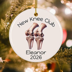 May include: A round white ceramic ornament featuring a gold hanging string. The text New Knee Club is printed in black above two metallic-toned anatomical knee joint illustrations. Below the graphics, the name Eleanor and the year 2026 are printed.